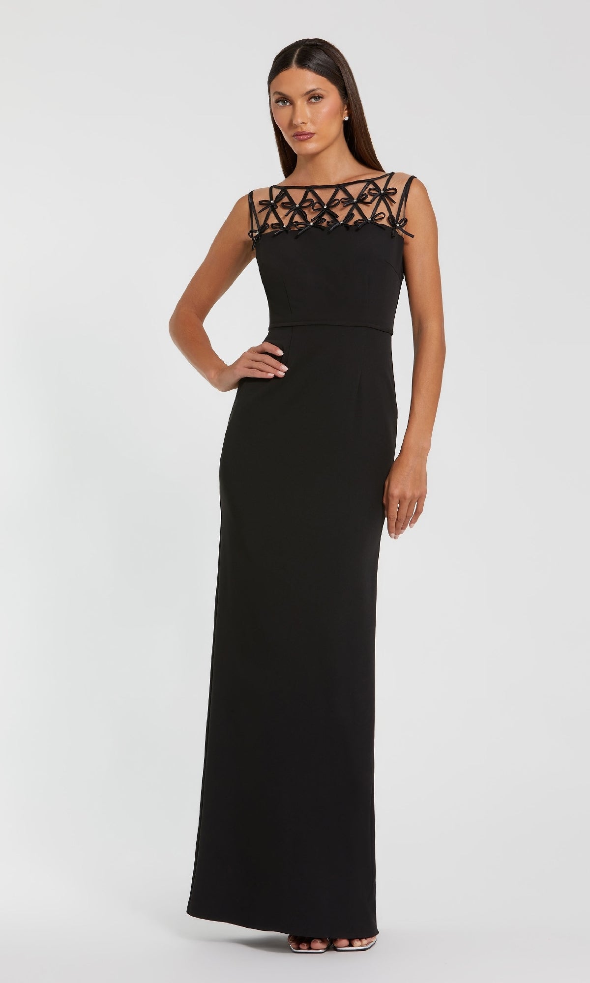 A woman models the Mac Duggal 2295 Bow-Neck Long Black Formal Dress with a sleeveless design and geometric sheer neckline, standing against a plain white background.